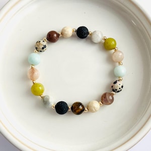 May include: A beaded bracelet featuring a variety of colorful gemstones, including brown, black, white, and green beads. The bracelet is strung on a gold-colored chain and is displayed on a white surface.