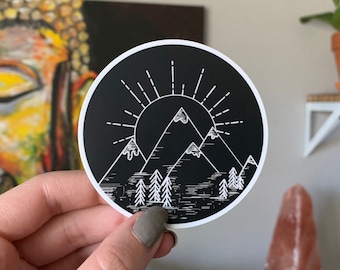 May include: A black and white sticker with a mountain range and sun design. The mountains are outlined in white and have a white sun with rays coming from it. The sticker is on a black background.