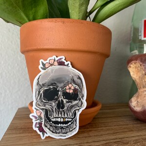 May include: A sticker of a gray skull with a single gold tooth, surrounded by pink and purple flowers. The skull has black eye sockets and a black nose.