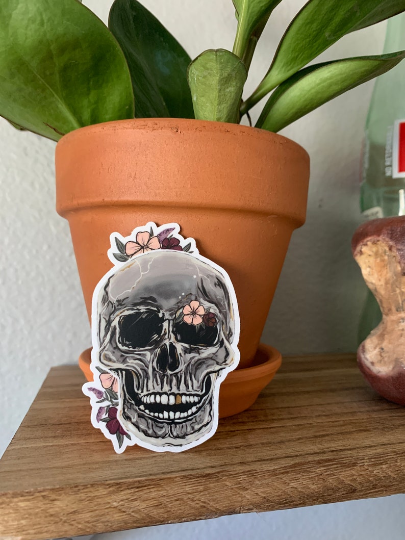 May include: A sticker of a gray skull with a single gold tooth, surrounded by pink and purple flowers. The skull has black eye sockets and a black nose.