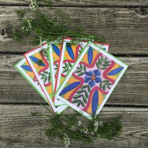 May include: A collection of colorful greeting cards with floral designs, featuring blue, orange, and green elements. The cards are arranged on a weathered wooden surface, with sprigs of greenery adding a natural touch.