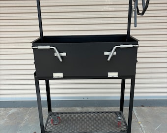May include: A black metal barbecue grill with a rectangular firebox, a warming rack, and a hand crank. The grill has a lower shelf and sits on four wheels with red rims. The background is a light brown roll-up door.