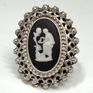 May include: Oval silver-toned ring with a black cameo featuring a white classical figure holding a plant. The ring has a detailed, openwork border with a scalloped edge. The cameo design is a classic, elegant style.