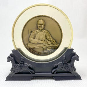 May include: A decorative bronze-toned plaque featuring a portrait of a man, set within a cream-colored circular frame. The plaque is mounted on a black base with stylized winged horse figures. The text "Dr. Sun Yat-Sen" is visible.