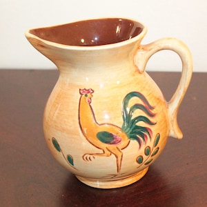 May include: A ceramic pitcher with a brown rim and a rooster design. The rooster is yellow with a green tail and red accents. The pitcher is decorated with green leaves.