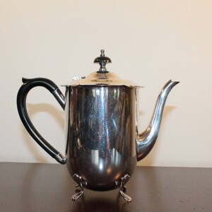 May include: A silver teapot with a black handle and three feet. The teapot has a shiny finish and a round body.