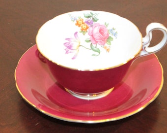 May include: A vintage teacup and saucer set with a floral design. The cup and saucer are both a deep burgundy red color with a gold trim. The floral design features a pink rose, purple tulips, and yellow and blue flowers.