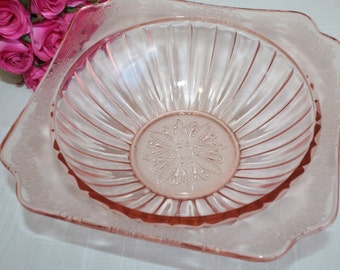 May include: A square pink glass bowl with a fluted design and a floral pattern in the center. The bowl is made of clear glass with a pink tint.