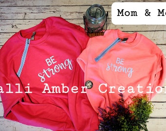 May include: Two pink sweatshirts with the text "Be strong" in white lettering. The sweatshirts are laid out on a wooden surface. The text "Mom & Me" is in a white box at the top of the image. The text "Calli Amber Creations" is at the bottom of the image.