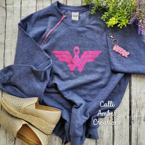 May include: A navy blue sweatshirt with a pink zipper and a pink Wonder Woman logo, paired with gold espadrille shoes and a keychain that says "WARRIOR". The sweatshirt has long sleeves.