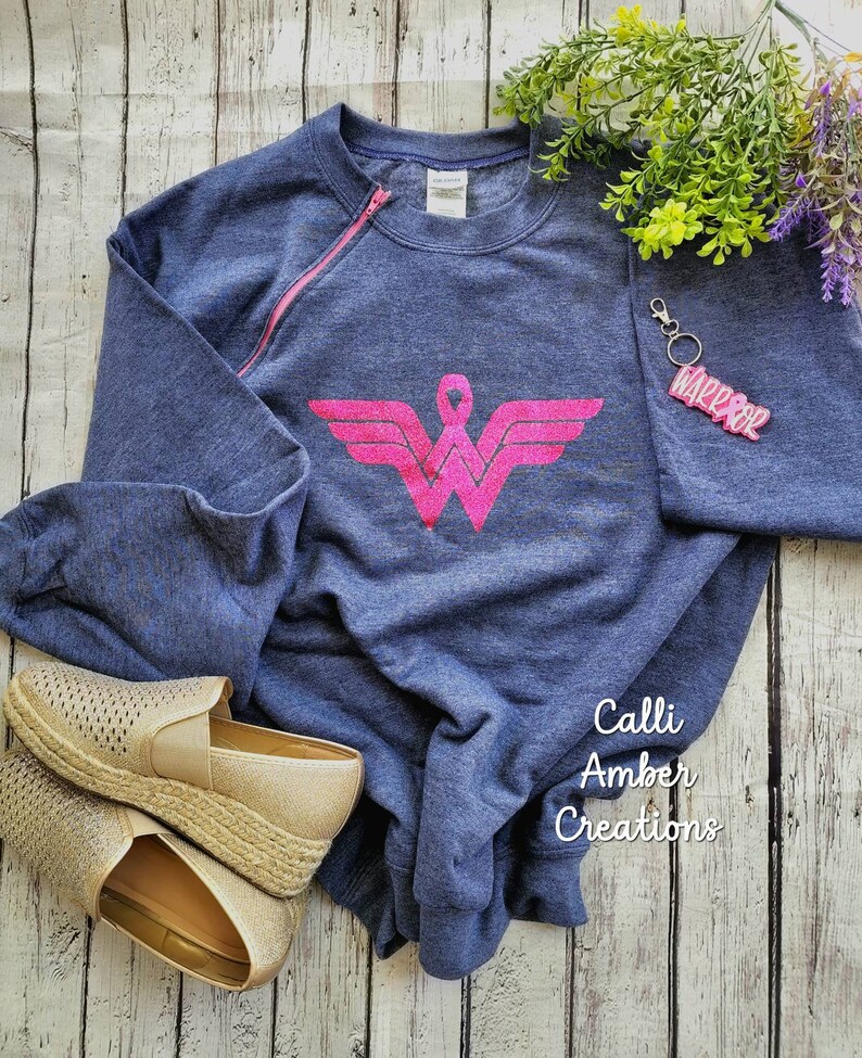 May include: A navy blue sweatshirt with a pink zipper and a pink Wonder Woman logo, paired with gold espadrille shoes and a keychain that says "WARRIOR". The sweatshirt has long sleeves.