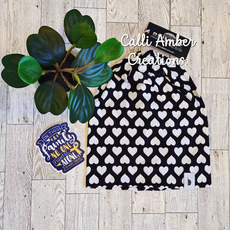 Op de afbeelding: Een zwarte muts met een herhalend wit hartjespatroon, een klein wit merklabel, een sticker met de tekst In This Family No One Fights Alone en een zwart visitekaartje met de tekst Calli Amber Creations in witte letters.