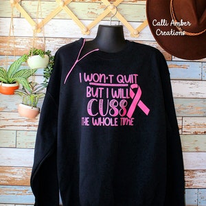 May include: Black sweatshirt with pink text that reads "I won't quit but I will cuss the whole time" and a pink ribbon. The sweatshirt is displayed on a black mannequin. A brown hat is visible in the background.