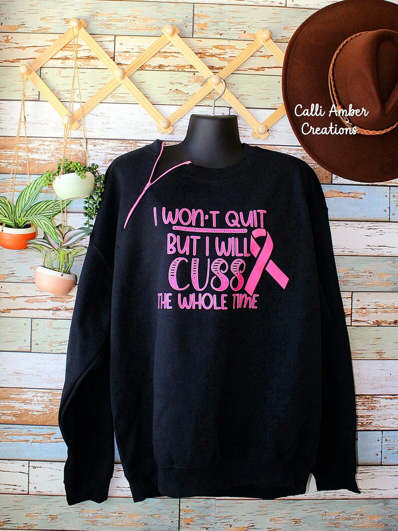 May include: Black sweatshirt with pink text that reads "I won't quit but I will cuss the whole time" and a pink ribbon. The sweatshirt is displayed on a black mannequin. A brown hat is visible in the background.