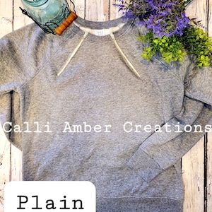 May include: A heather gray sweatshirt with a crew neck and long sleeves. The sweatshirt has white drawstrings and the text "Calli Amber Creations" and "Plain". The background includes a mason jar, purple flowers, and green foliage.