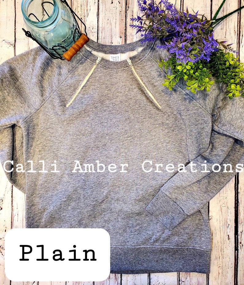 May include: A heather gray sweatshirt with a crew neck and long sleeves. The sweatshirt has white drawstrings and the text "Calli Amber Creations" and "Plain". The background includes a mason jar, purple flowers, and green foliage.