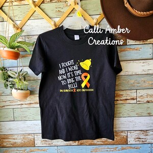 May include: Black t-shirt featuring a yellow and orange ribbon, a bell, and the text "I fought and I won! Now it's time to ring the bell!" with the dates "DX: 12/18/2018 EOT: 08/01/2024". The shirt is hanging on a wooden rack with a brown hat on the right side.