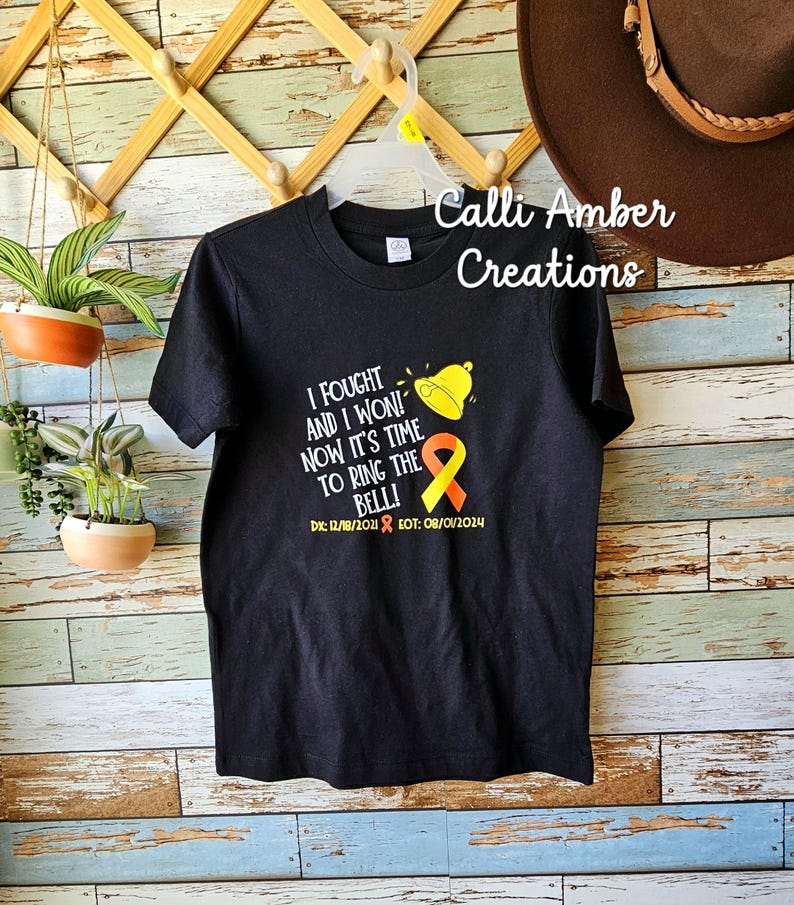 May include: Black t-shirt featuring a yellow and orange ribbon, a bell, and the text "I fought and I won! Now it's time to ring the bell!" with the dates "DX: 12/18/2018 EOT: 08/01/2024". The shirt is hanging on a wooden rack with a brown hat on the right side.