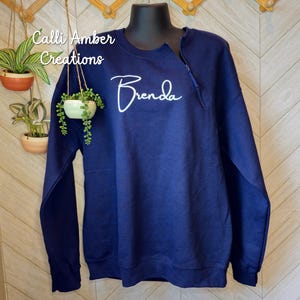 May include: A navy blue sweatshirt with a zipper detail at the neckline. The name "Brenda" is written in white script across the chest. The text "Calli Amber Creations" is in the upper left corner.