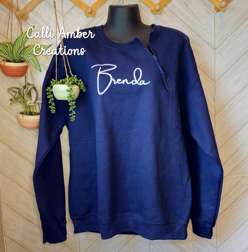 May include: A navy blue sweatshirt with a zipper detail at the neckline. The name "Brenda" is written in white script across the chest. The text "Calli Amber Creations" is in the upper left corner.
