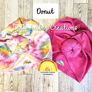 Op de afbeelding: Twee geknoopte hoofdbanden van Calli Amber Creations. De ene heeft een veelkleurig tie-dye patroon, terwijl de andere effen felroze is met witte spikkels. Een kleine sticker ertussen leest More Precious Than Gold Childhood Cancer Awareness.