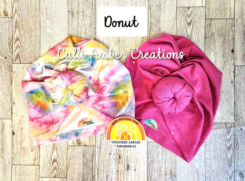 Op de afbeelding: Twee geknoopte hoofdbanden van Calli Amber Creations. De ene heeft een veelkleurig tie-dye patroon, terwijl de andere effen felroze is met witte spikkels. Een kleine sticker ertussen leest More Precious Than Gold Childhood Cancer Awareness.