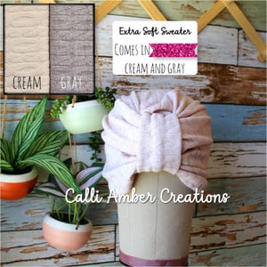 Op de afbeelding: Een lichtroze gebreide tulband met een centraal knoopdetail, weergegeven op een etalagepophoofd. De tekst op de afbeelding luidt: Extra Soft Sweater, Comes in, Cream and Gray en Calli Amber Creations. De stof heeft een subtiel textuurpatroon.