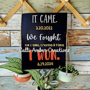 May include: Black chalkboard sign with white text that reads "IT CAME. 3.20.2022 We fought FOR 2 YEARS, 3 MONTHS & 9 DAYS Calli Amber Creations I WON. 6.29.2024" hanging from a wooden rack with three potted plants.