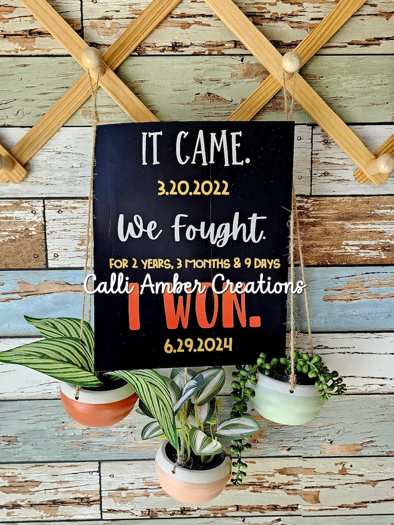 May include: Black chalkboard sign with white text that reads "IT CAME. 3.20.2022 We fought FOR 2 YEARS, 3 MONTHS & 9 DAYS Calli Amber Creations I WON. 6.29.2024" hanging from a wooden rack with three potted plants.