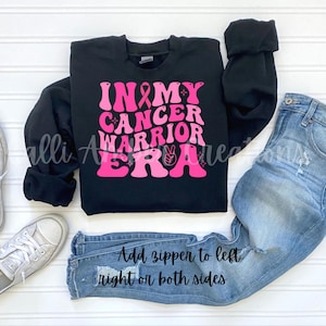 May include: Black sweatshirt with the text "IN MY CANCER WARRIOR ERA" in pink lettering, a pink ribbon, and a peace sign. The sweatshirt is paired with light blue jeans and silver sneakers. Text on the jeans reads "Add zipper to left right or both sides."