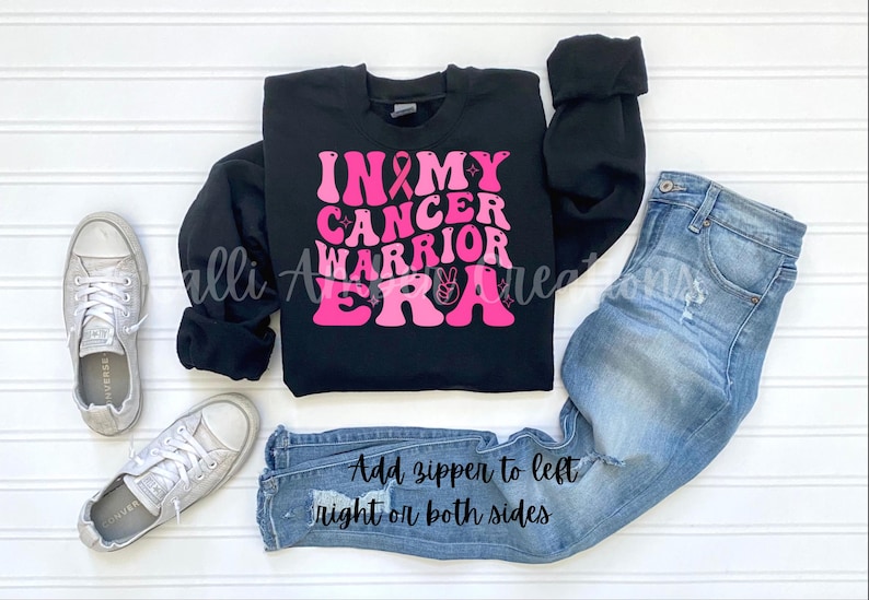 May include: Black sweatshirt with the text "IN MY CANCER WARRIOR ERA" in pink lettering, a pink ribbon, and a peace sign. The sweatshirt is paired with light blue jeans and silver sneakers. Text on the jeans reads "Add zipper to left right or both sides."