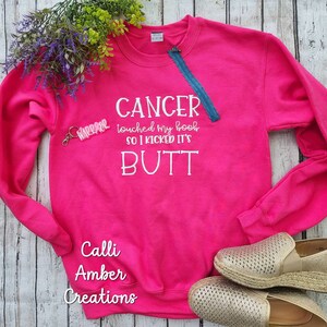May include: A bright pink sweatshirt with the text "CANCER touched my boob SO I KICKED IT'S BUTT" in white. A small "WARRIOR" keychain and a blue zipper detail the design. The sweatshirt is paired with gold espadrille shoes.