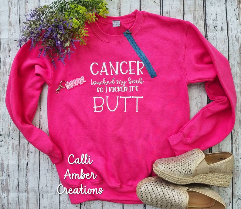 May include: A bright pink sweatshirt with the text "CANCER touched my boob SO I KICKED IT'S BUTT" in white. A small "WARRIOR" keychain and a blue zipper detail the design. The sweatshirt is paired with gold espadrille shoes.