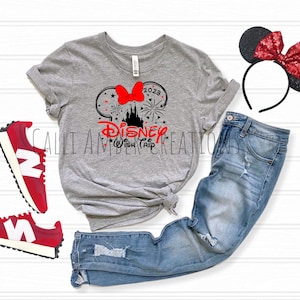 May include: A gray t-shirt with a black and red graphic of Mickey Mouse ears with a bow, fireworks, and the text "Disney Wish Trip 2023". The shirt is paired with blue jeans, red sneakers, and a black headband with red glitter ears.