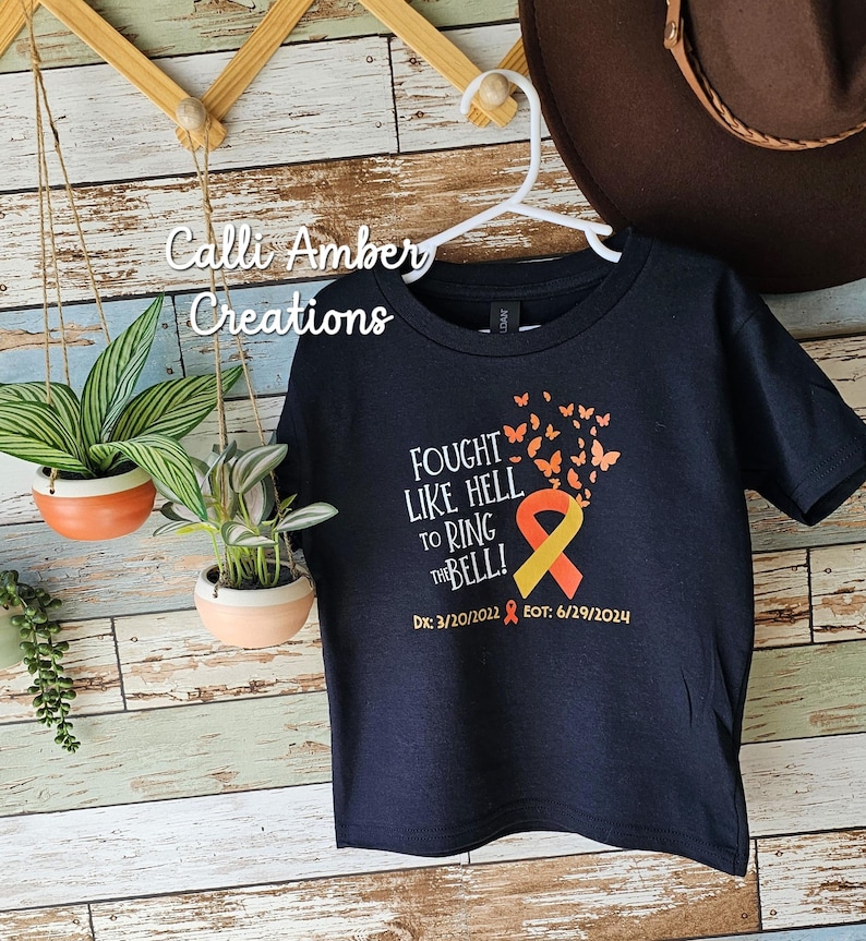May include: Black t-shirt with a yellow and orange ribbon and butterflies. The text reads "Fought Like Hell To Ring The Bell!" with the dates "Dx: 3/20/2022 EOT: 6/29/2024".