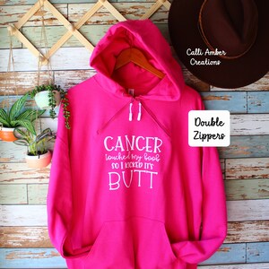 May include: A bright pink hoodie with a front pocket and double zippers. The hoodie has white text that reads "CANCER touched my boob so I kicked its BUTT". A brown hat hangs in the background.
