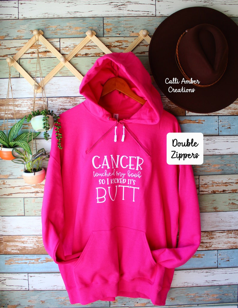 May include: A bright pink hoodie with a front pocket and double zippers. The hoodie has white text that reads "CANCER touched my boob so I kicked its BUTT". A brown hat hangs in the background.