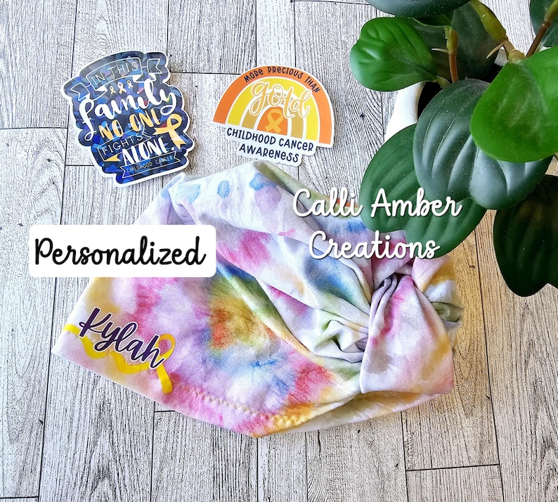 Op de afbeelding: Een tie-dye stoffen hoofdband met de naam Kylah in paars schrift en een gele lintafbeelding. Twee stickers zijn inbegrepen: een met de tekst In this family no one fights alone en een met een regenboog en de tekst More precious than gold.