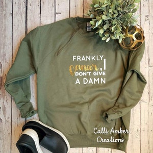 May include: Olive green sweatshirt with the text "Frankly, I don't give a damn" in white and gold glitter lettering. Black slip-on shoes with white soles are at the bottom left. A small green plant and gold decorative ball are at the top right.