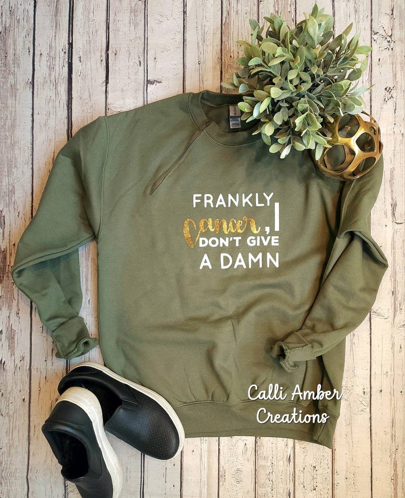 May include: Olive green sweatshirt with the text "Frankly, I don't give a damn" in white and gold glitter lettering. Black slip-on shoes with white soles are at the bottom left. A small green plant and gold decorative ball are at the top right.