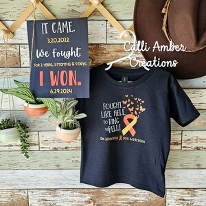 May include: A black t-shirt with a yellow and orange ribbon and butterflies printed on the front. The text on the shirt reads "Fought Like Hell To Ring The Bell!" with the dates "Dx: 3/20/2022 EOT: 6/29/2024".