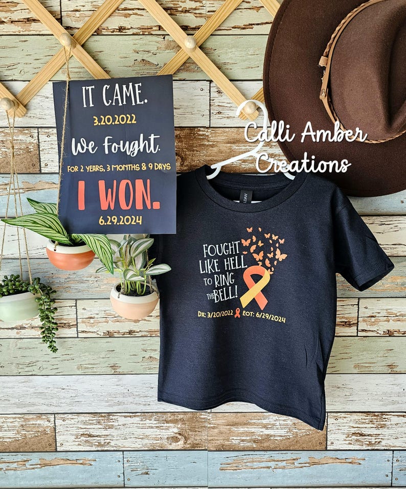 May include: A black t-shirt with a yellow and orange ribbon and butterflies printed on the front. The text on the shirt reads "Fought Like Hell To Ring The Bell!" with the dates "Dx: 3/20/2022 EOT: 6/29/2024".