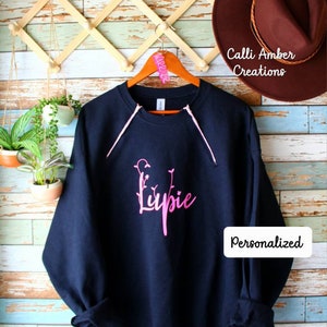 May include: Navy blue sweatshirt with pink accents and the word "Lupie" in a floral font. The sweatshirt is hanging on a wooden hanger. The word "Personalized" is in a white rectangle on the sweatshirt.