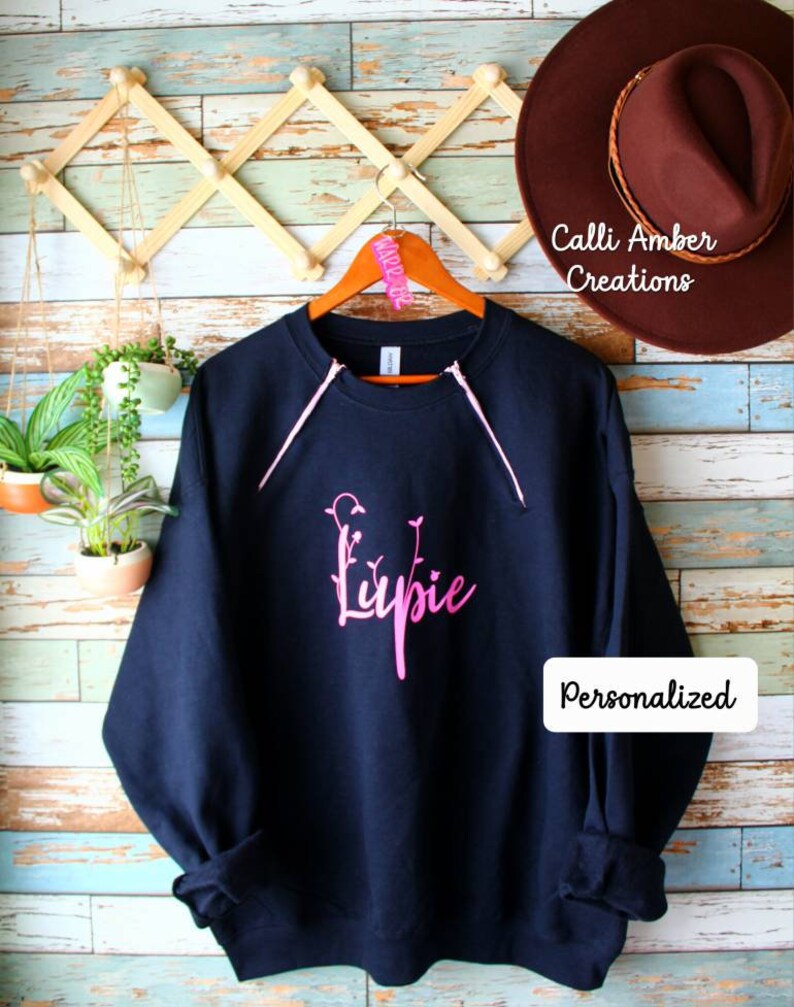 May include: Navy blue sweatshirt with pink accents and the word "Lupie" in a floral font. The sweatshirt is hanging on a wooden hanger. The word "Personalized" is in a white rectangle on the sweatshirt.