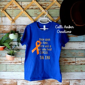 May include: A royal blue t-shirt with an orange ribbon and the text "Once upon a time, there was a boy who beat CANCER... THE END". The shirt is hanging on a white hanger against a wooden background.