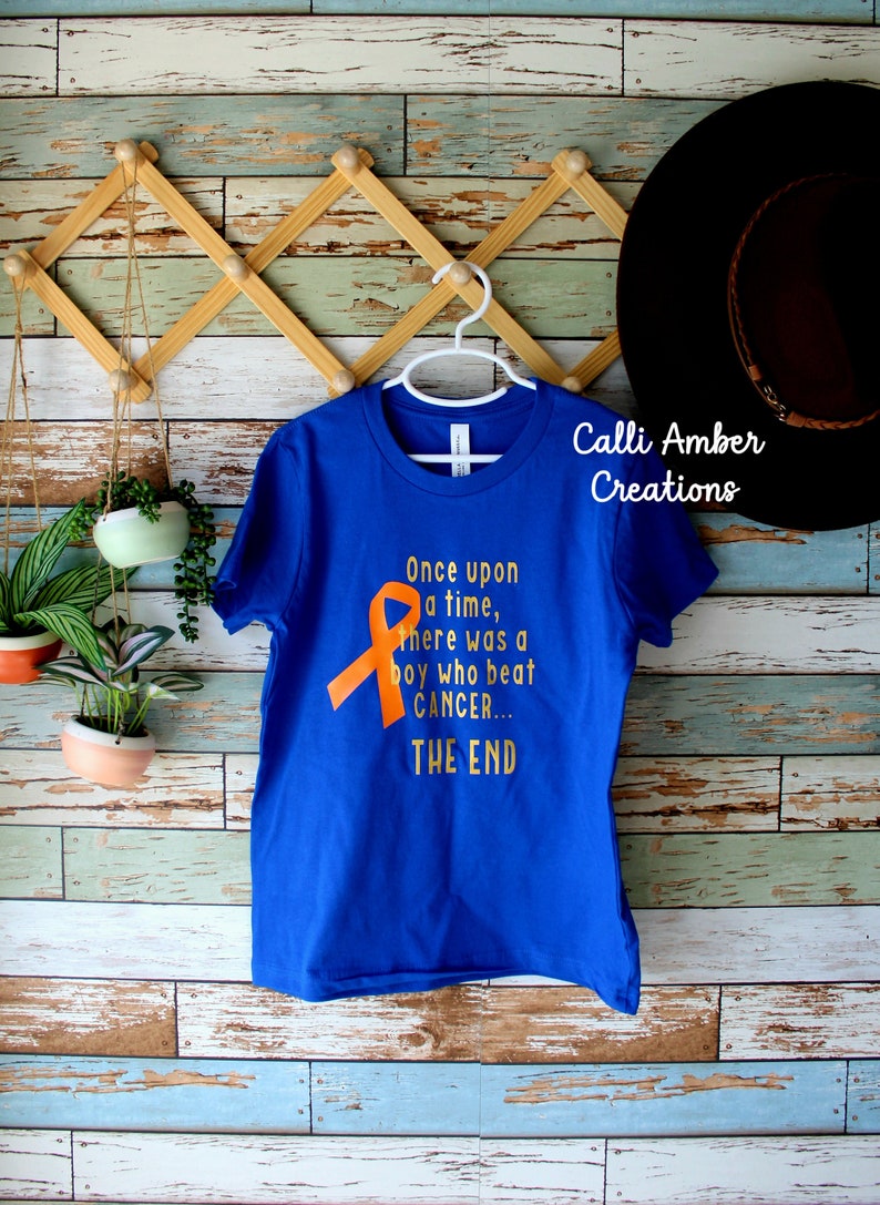 May include: A royal blue t-shirt with an orange ribbon and the text "Once upon a time, there was a boy who beat CANCER... THE END". The shirt is hanging on a white hanger against a wooden background.
