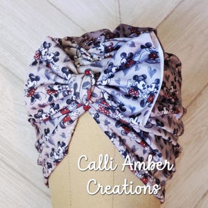 Op de afbeelding: Een babyhoofdband in tulbandstijl met een Mickey Mouse-print in rood, zwart en wit op een grijze achtergrond. De hoofdband heeft een grote strik en de witte tekst "Calli Amber Creations".