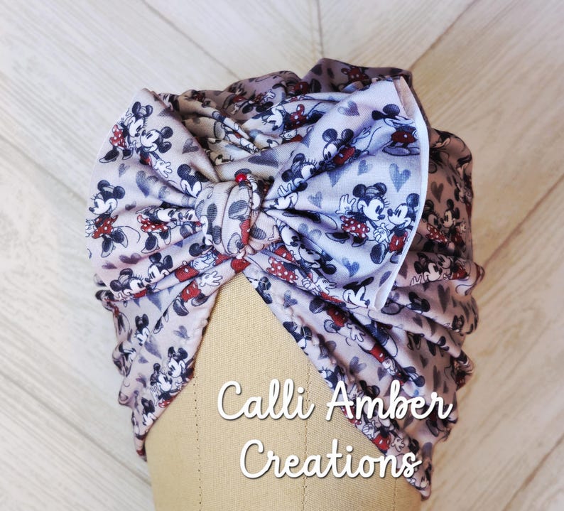 Puede incluir: Un turbante de color claro con un patr&oacute;n repetitivo de personajes de Mickey y Minnie Mouse y corazones peque&ntilde;os. La tela est&aacute; fruncida en la parte delantera formando un lazo grande y prominente. El texto Calli Amber Creations es visible.