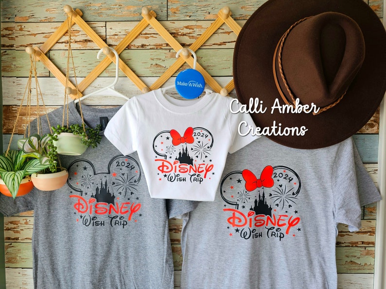 May include: Three gray and white t-shirts with a black and red Mickey Mouse ears design. The shirts have the text "2024 Disney Wish Trip" on them. The shirts are hanging on a wooden rack with a brown hat on top.