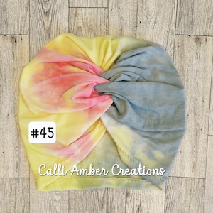 Op de afbeelding: Een tie-dye tulband met een gedraaid ontwerp aan de voorzijde in gemengde kleuren geel, roze en blauw. De stof heeft een zacht, gem&ecirc;leerd patroon. Een wit vierkant label aan de linkerkant bevat de tekst #45 en Calli Amber Creations.